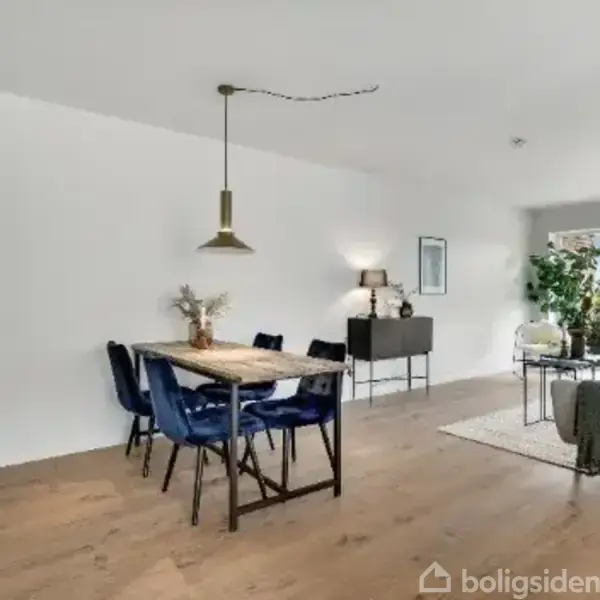 En spisestue med et træbord og blå stole, belyst af en hængelampe. I baggrunden ses en stue med sofa, gulvtæppe og adgang til en balkon gennem glasdøre.