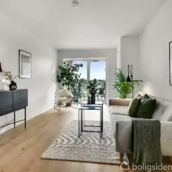 En stue med en grå sofa til højre, der vender mod et sofabord. Et mørkt skab til venstre ved siden af en lampe. Store vinduer bagved giver udsigt til en altan. En plante står ved døren.