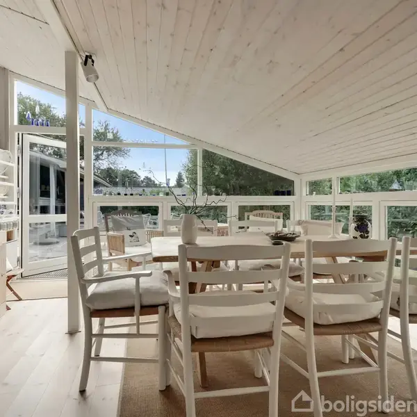 Spisebord med hvide stole er placeret i et lyst, rummeligt rum med store vinduer og skrå lofter, der kigger ud på grønne omgivelser. Møblerne er lyse og minimalistiske.