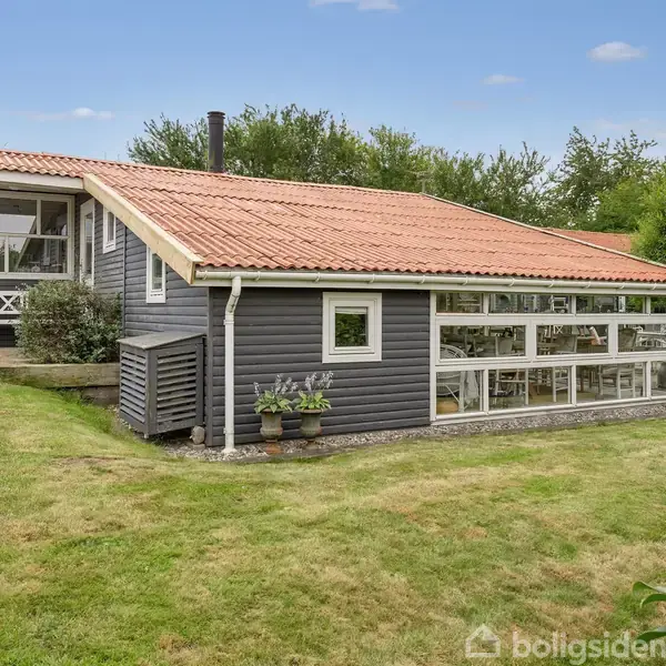Et sort træhus med rødt tag står i en grøn have. Store vinduer langs siden indrammer en indbydende udestue. Omkring huset er der frodige buske og græsplæne.