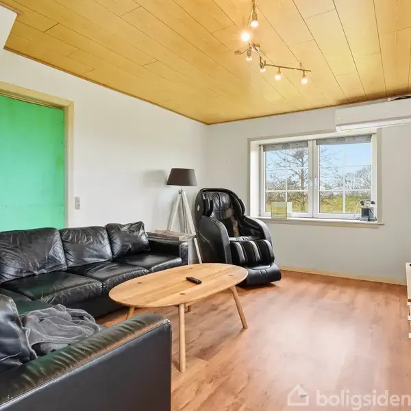 Sort lædersofa og massage-stol står i en stue med trægulv og træloft. Et kaffebord og en gulvlampe supplerer rummet, der har et vindue med udsigt til træer.