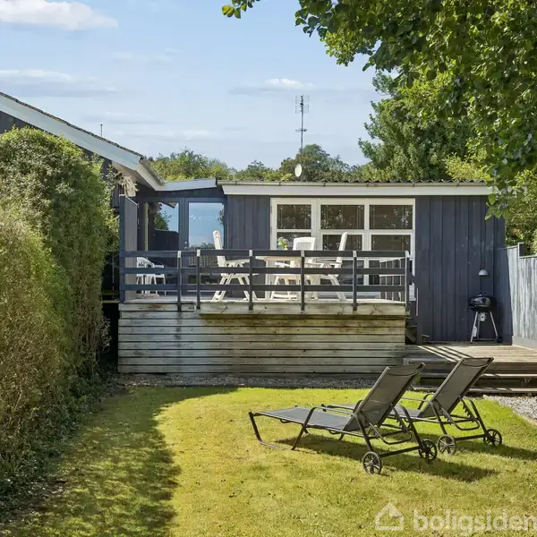 Sommerhus med træterrasse er omgivet af hække. To liggestole står på græsplænen. I baggrunden ses blå himmel og enkelte skyer.