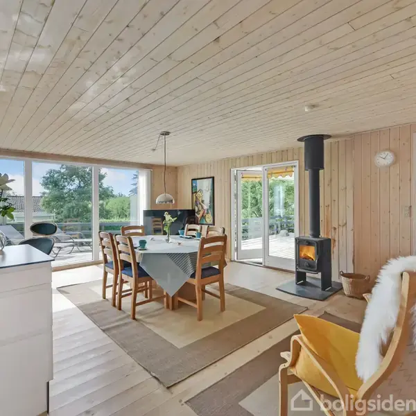 Et moderne køkken-alrum med spisebord, træloft og brændeovn. Store vinduer afslører en grøn have. En lampe hænger over bordet, og der er en dør til terrassen.