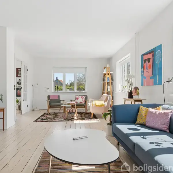 Stue med blå sofa og farverige puder på et tæppe. To stole ved et bord står nær vinduer, der slipper lys ind. Vægdekorationer og planter i en lys, moderne atmosfære.