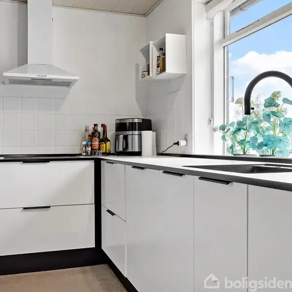 Et moderne køkken med hvide skabe og en sort vandhane. Flasker og en kaffemaskine står på bordpladen. Et vindue med udsigt lukker sollys ind.