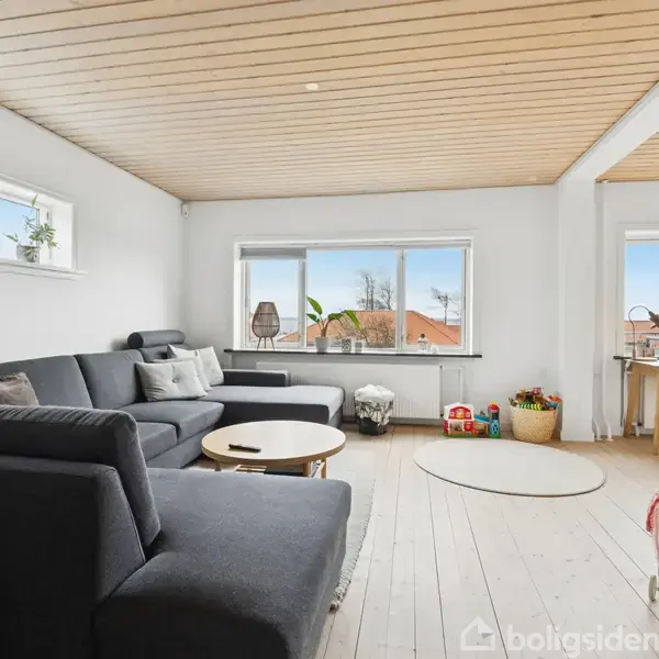 En stor, mørkegrå sofa står i en lys stue med trægulv og træloft. Store vinduer giver udsigt til hustage. En rund træbord og nogle potteplanter ses i rummet.