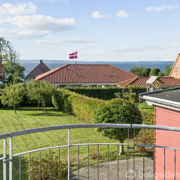En balkon med gelænder og udsigt over en velplejet have. I baggrunden ses huse med rødt tag og en dansk flag. Havet er synligt under en blå himmel.