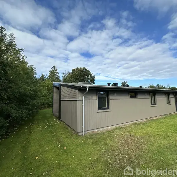 Et enkelt, rektangulært hus står på en grøn græsplæne, omgivet af træer under en delvist skyet himmel.