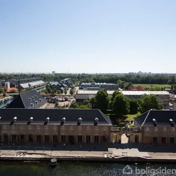 Bygninger med mørke tage ligger langs en kanal. I baggrunden ses grønne træer og andre bygninger. Himlen er lys og klar, og vandet reflekterer omgivelserne.