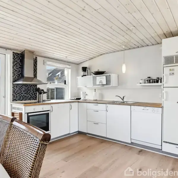 Et lyst, moderne køkken med hvidt inventar og træloft. Køkkenet indeholder en ovn, emhætte, og køleskab. Spisebord med flettede stole ses forrest. Store vinduer giver udsigt til haven.
