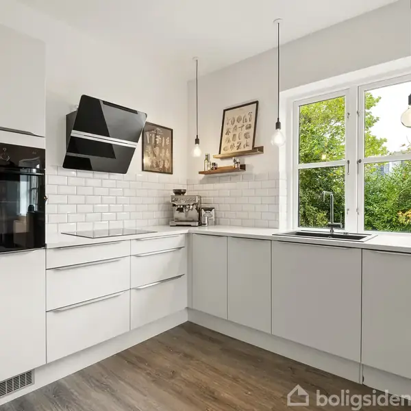 Et moderne køkken med hvide skabe og en indbygget ovn. Vindue giver udsigt til træer udenfor. Hængende lamper og kaffemaskine på bordpladen tilføjer funktionalitet.