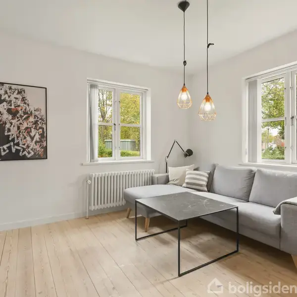 Gråt hjørnesofa står i lys stue med to vinduer. Loftslampen hænger over sofabordet. Radiator under vindue og abstrakt kunstværk på væggen. Trægulve og neutral vægfarve.