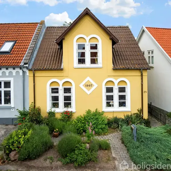 Gult hus med buede vinduer, dekoreret med tallet "1907", omgivet af have og beliggende mellem grå og hvidt hus. Huset har en brun tagryg og en frodig forhave.