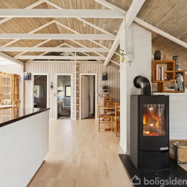 Brændeovn opvarmer stue med trævægge og loftsbjælker, omgivet af spiseområder og indgang. Hyggelig atmosfære skabes af varme træelementer og en brændende flamme, set gennem glasset.