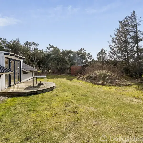Et moderne sommerhus med sort tag står på græsplæne ved siden af en træterrasse. Omkring huset er der træer og buske i et naturpræget landskab.