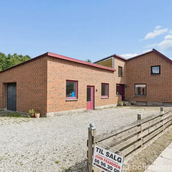 En moderne, rød murstensbygning med fladt og skråt tag står stille på grusbelagt grund ved siden af en fortovssti. Et skilt foran bygningen siger "TIL SALG, John Frandsen, 86 96 30 00".