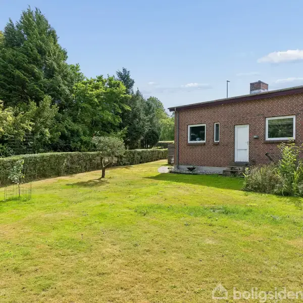 En murstensbygning står ved en græsklædt have med en hækkeomkranset have. Bygningen har to vinduer og en dør. I baggrunden ses frodig vegetation og træer.