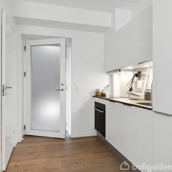 Et moderne køkken har hvide skabe og skuffer, en indbygget ovn, og en vask på bordpladen. Rummet er lyst med en glasdør, der lader naturligt lys strømme ind.