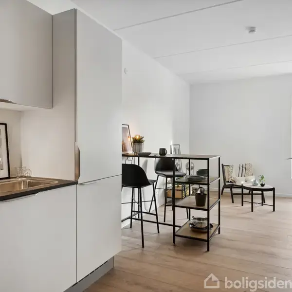 Et moderne køkken med hvide skabe og en vask på en træbordplade. Rummet omfatter en lille spiseplads med høje stole og et reolbord, badet i naturligt lys fra et vindue.