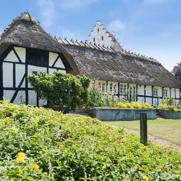 Stråtækt bindingsværkshus ligger i en blomstrende have på en solrig dag. Huset er stort med hvide og sorte vægge, og omgivet af grønne buske og blomsterbede.