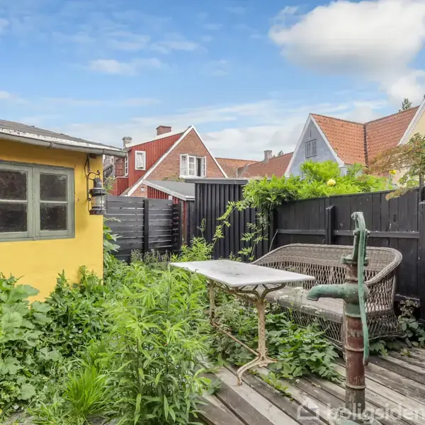 En gammel havebænk og et bord står i en lille, tilgroet have ved siden af et gult hus. Sort hegn omslutter området, og der ses nabohuse i baggrunden.