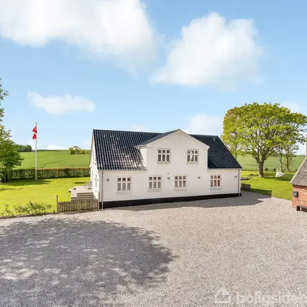 Hvidt hus står stille på en grusbelagt plads, omgivet af grønne marker og træer. Der er en flagstang med et dansk flag i haven.