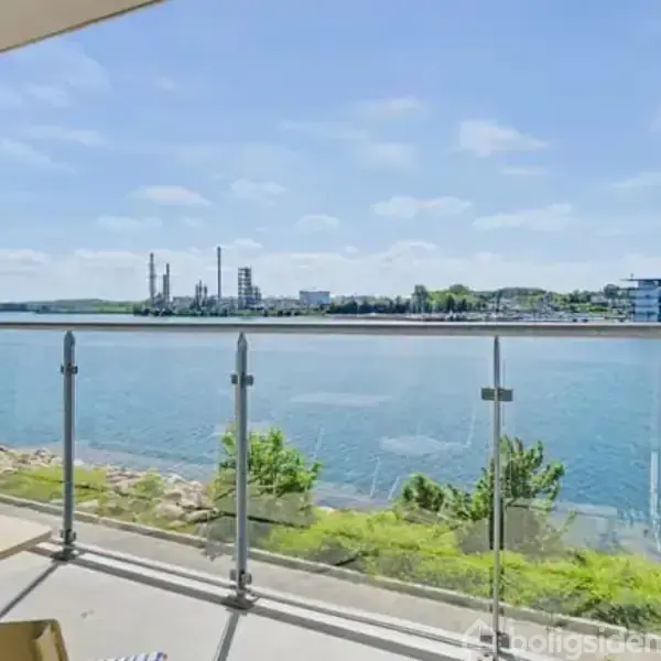 Altan med glasrækværk har udsigt over en flod. I baggrunden ses industrielle bygninger og moderne lejlighedsbygninger, omgivet af grønne områder under en blå himmel.
