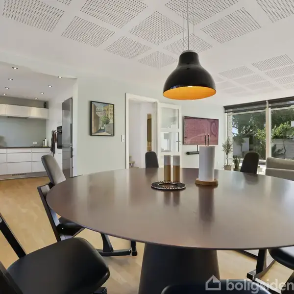 Rundt spisebord med sorte stole omkring, under en hængende lampe. Baggrunden viser et moderne køkken og stue med store vinduer, der bringer naturligt lys ind.