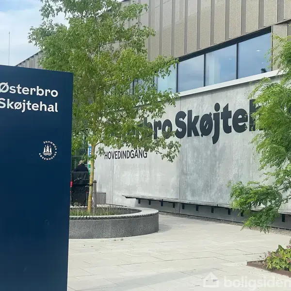 Skiltet viser "Østerbro Skøjtehal" og står foran en moderne bygning med teksten "HOVEDINDGANG" på væggen. Omkring er der træer og cykler.