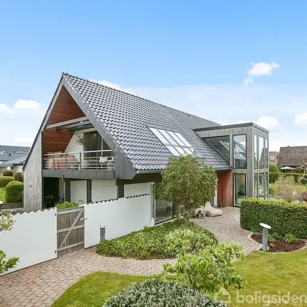 Et moderne hus med skråt tag står i en velplejet have. Stor glasfacade og altan tilføjer elegance. Beliggende i et roligt boligområde med andre lignende huse i baggrunden.