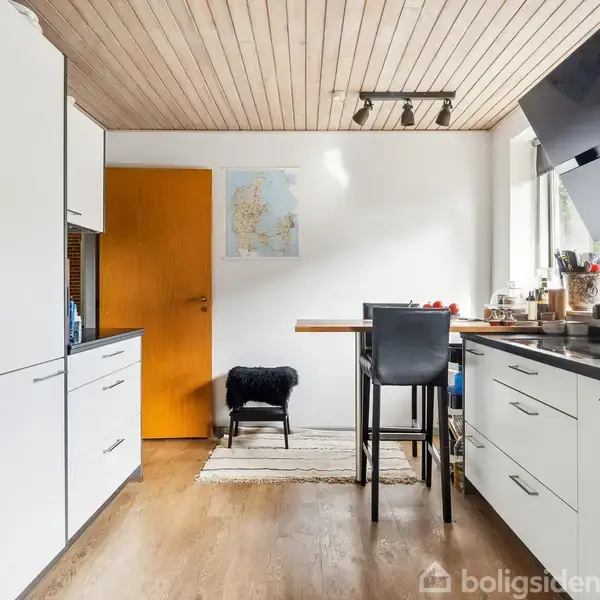 Et moderne køkken viser hvidt skabsinventar, stålovne, og en emhætte. Et lille bord med stole står i midten. Kort på væggen og dør i baggrunden, træloft og gulv.