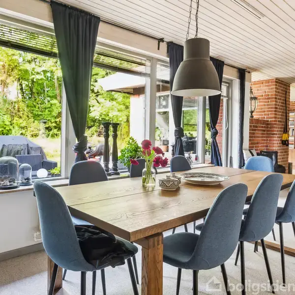 Langt spisebord med seks blå stole omkring det. Bordet er dekoreret med blomster og tallerkener. Store vinduer med mørke gardiner giver udsigt til en grøn have.
