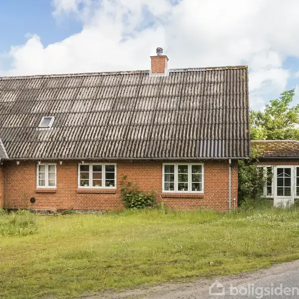Et rødt murstenshus med skråt tag står på en græsklædt grund. Huset har flere vinduer og en skorsten. Himmel med spredte skyer ses i baggrunden.