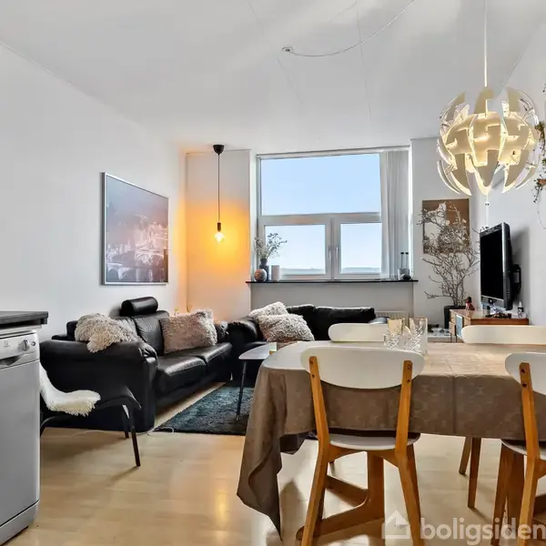 En moderne stue med sort lædersofa, placeret ved et stort vindue, hvor lys kommer ind. Der er et spisebord med træstole; en dekorativ lampe hænger over bordet.