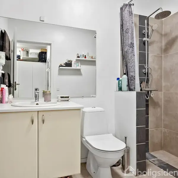 Badeværelse med vaskeskab og spejl, toilet, og brusekabine med gråt flisegulv. Hvide vægge og hylde med toiletartikler i baggrunden. En dør fører ud fra rummet.