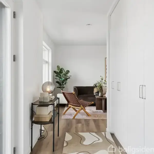 En lys gang med hvide skabe langs højre side fører til en stue. Et bord med en lampe og bøger står til venstre. Stuen har en brun stol og grønne planter.