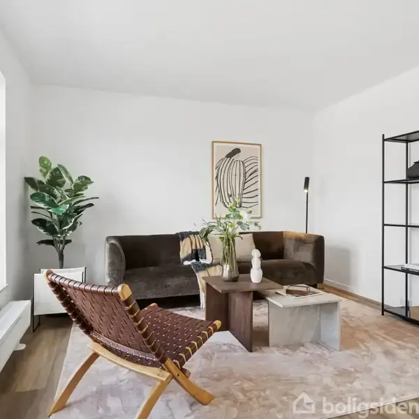 Stue med moderne møbler: en brun sofa, et vævet lænestol og et sofabord. Hylde med planter og bøger i baggrunden. Store vinduer giver naturligt lys.