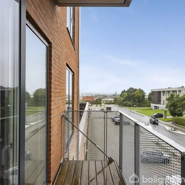 En balkon med metalrækværk og trægulv strækker sig fra en rød murstensbygning, med udsigt over en vej, træer og moderne bygninger i baggrunden.