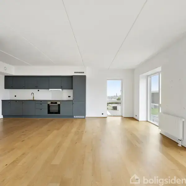 En lys, rummelig stue med trægulv og et moderne køkken med blå skabe. Rummet har store vinduer, der giver udsigt til en balkon og udenfor. Ingen tekst til stede.