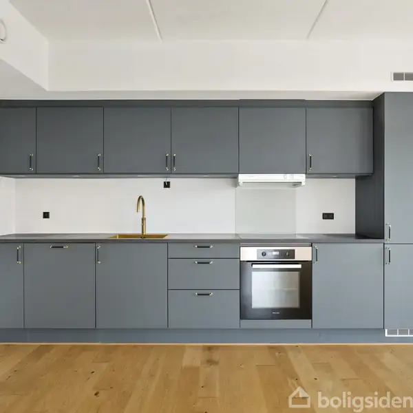Køkken med grå skabe, træbordplade og åbent rum. Indbygget ovn og vask ses. Skabene er i moderne design mod en hvid væg med trægulv.