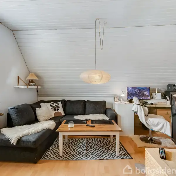 En mørkegrå sofagruppe med lammeskindstæpper står i en stue under en skråning. Sofabordet har en træplade, og der hænger en lampeskærm over det. I baggrunden ses et skrivebord med computer udstyr.