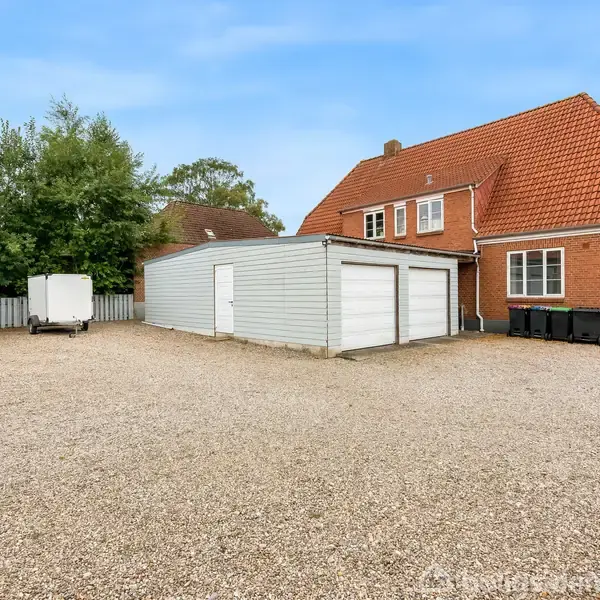 Rødt murstenshus med rødt tag står ved grusplads, flankeret af grå garagebygninger. Pladsen har en parkeret hvid trailer. Træer og hegn udgør baggrunden under en blå himmel.