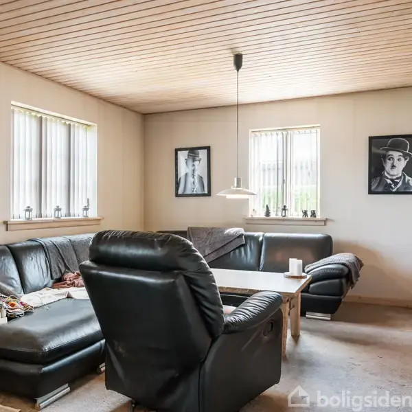 En stue med en sort lædersofa og lænestol står omkring et træbord. To billeder hænger på væggen, og vinduer giver naturligt lys. En lampe hænger fra loftet.
