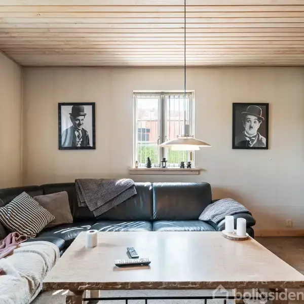 En sort lædersofa med tæpper og puder står langs en væg. Foran står et sofabord med en fjernbetjening og en kop. To portrætter hænger på væggen.