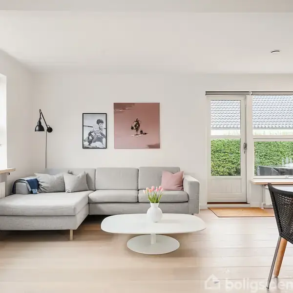 En lys stue med en grå hjørnesofa og et rundt sofabord med en vase. Der er to billeder på væggen. Store vinduer og en glastør fører ud til en terrasse.