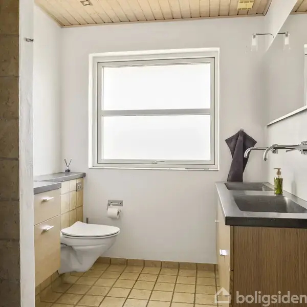 Badeværelse med toilet hængende på væggen, håndvask med armatur, spejl, bruser, lys beige fliser på gulvet, og stort vindue. Hvide vægge og træloft skaber et lyst rum.