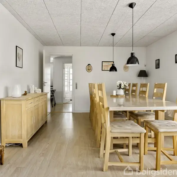 Et langt spisebord af træ med otte stole er centreret i et lyst spisestuerum. Et vitrineskab står til venstre ved væggen, dekoreret med små figurer.