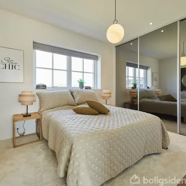 Soveværelse med dobbeltseng dækket af lys sengetæppe og puder, reflekteres i store skabspejle. Natbord med lampe ved siden, og væggen har teksten "Stay CHIC".