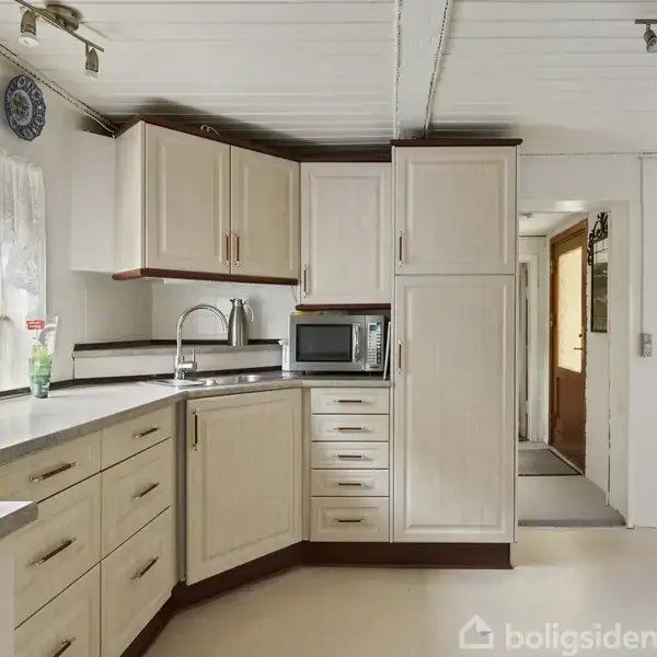Køkken med lyse træskabe og skuffer, hvor en mikrobølgeovn er placeret på bordet. En vask er monteret under et vindue med hvide gardiner. En gang fører til en dør.