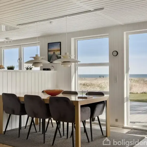 Spisebord står dækket af lamper og omgivet af stole i et lyst rum. Store vinduer og en glasdør viser en udsigt til stranden og havet udenfor.